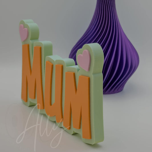 Side view of a 3D printed Mum nameplate with heart-shaped accents on top of each letter. The layered design shows the yellow-blue base, bold orange letters, and pink hearts, creating a vibrant, freestanding display. Ideal for a personalised Mothers Day gift, birthday gift or home decor.